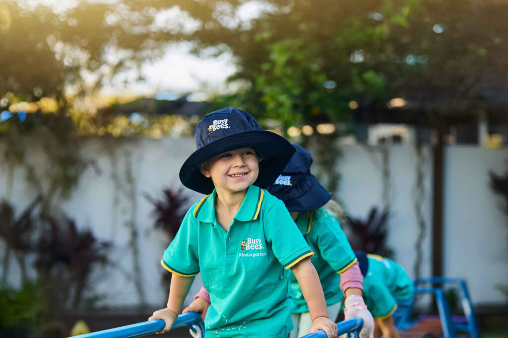 Young boy in Busy Bees teal sun safe polo shirt and Busy Bees wide brim hat smiling on playground equipment during Outdoor Play