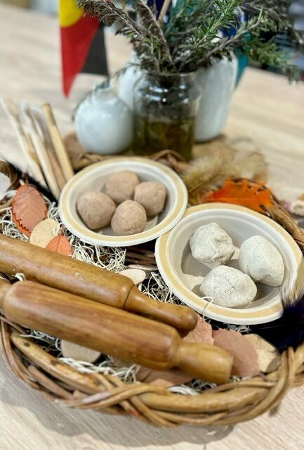 A wicker tray holds two bowls of round objects, wooden rolling pins, dried leaves, and a jar with greenery.
