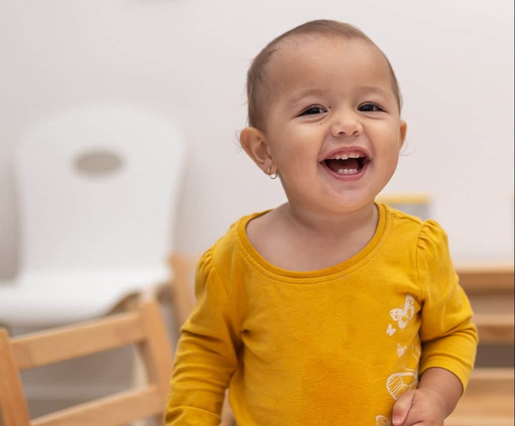 A toddler in a yellow shirt with butterfly designs smiles and looks at the camera while standing indoors, capturing a moment of early learning joy. Furniture is visible in the background, creating a warm and inviting childcare environment.