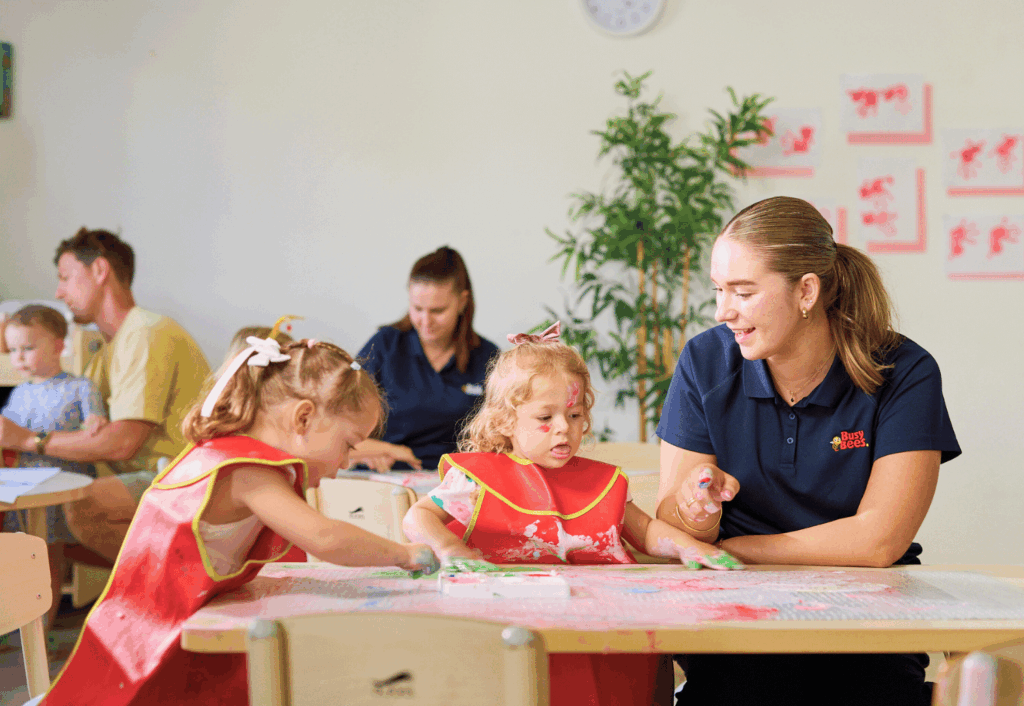 Young children wearing aprons paint at a table with a teacher assisting; other adults and children work in the background in a classroom setting.