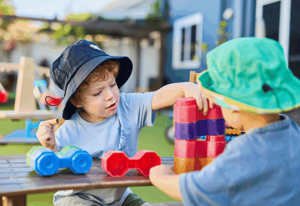 Two young children wearing hats play with colorful plastic blocks and toys on a wooden table outdoors in a sunny yard.
