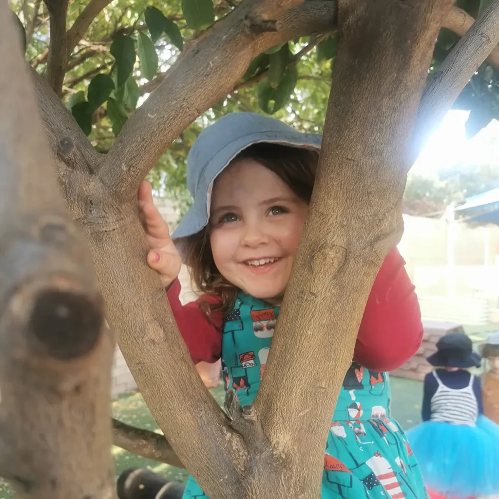 A young child wearing a teal dress and a gray sun hat smiles while playing in a tree. Other children are visible in the background.