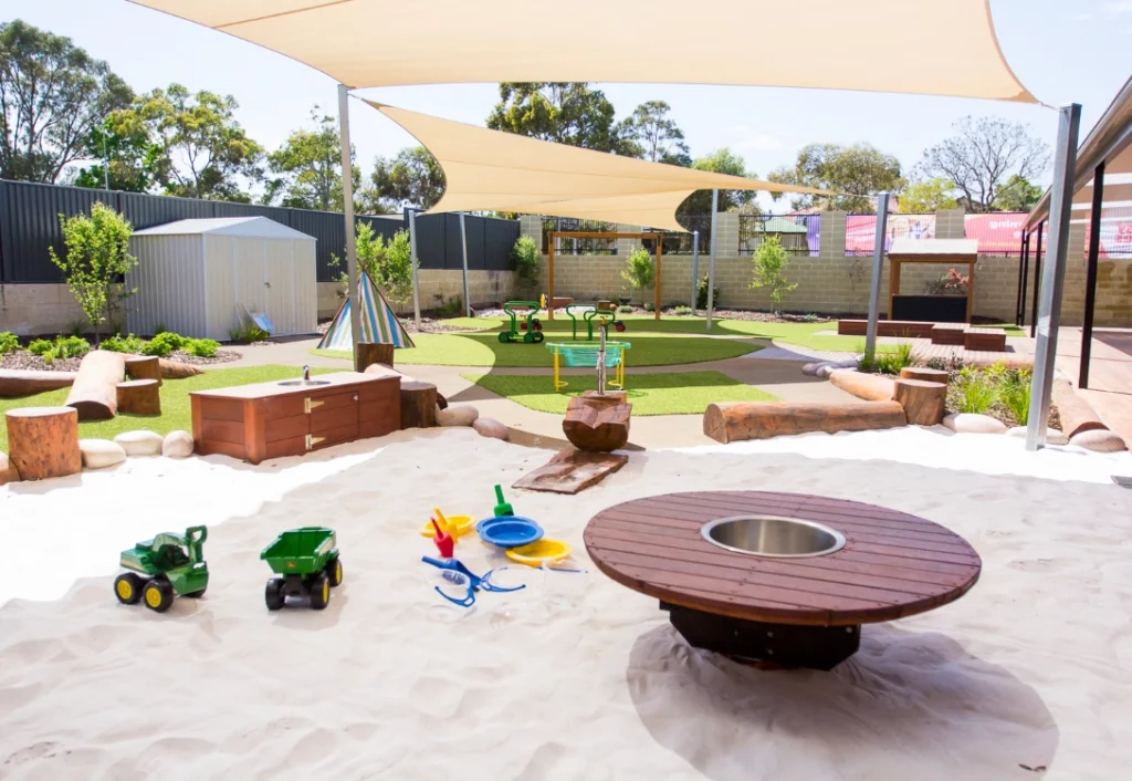 A children's outdoor play area with a sandpit, toy trucks, colorful sand toys, a shaded seating area, and various playground equipment on artificial grass. Trees and a fence surround the space.