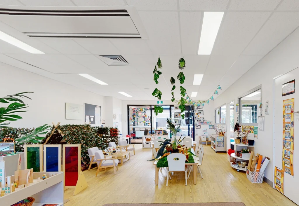 A brightly lit, organized classroom is adorned with educational materials, colorful decorations, and child-sized tables and chairs. Green plants hang from the ceiling, adding a touch of nature.