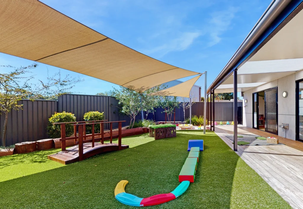Outdoor play area with artificial grass, a small wooden bridge, colorful play structures, and shade sails above. The area is adjacent to a building with a covered porch.