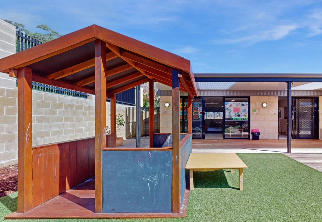 Playground area with a small wooden playhouse and a low table on artificial grass, in front of a building with large windows and doors.