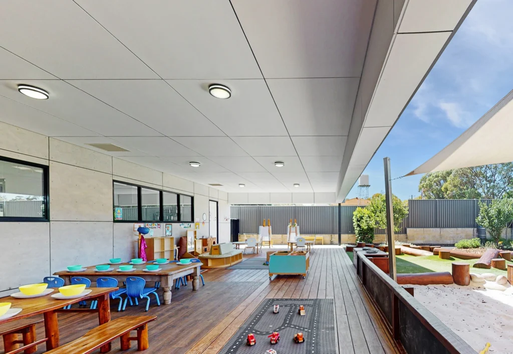 A childcare facility with a covered outdoor play area featuring tables, chairs, toys, and learning materials. There is also a sandbox and a mini-golf section.