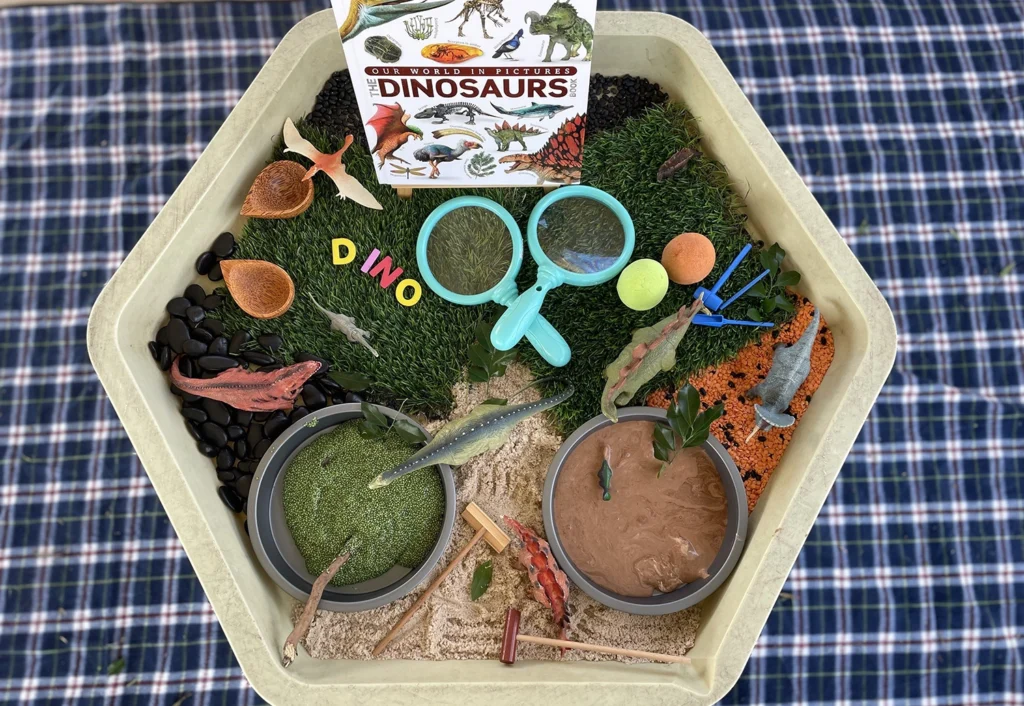 A sensory bin featuring dinosaur-themed items, including plastic dinosaurs, a magnifying glass, a book titled "Dinosaurs," and various textured materials like sand, beans, and moss.