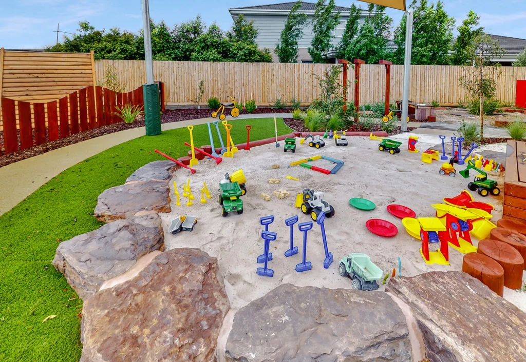 A colorful children's playground with a large sandpit filled with various toys, including shovels, trucks, and buckets. The play area is surrounded by greenery and a wooden fence.