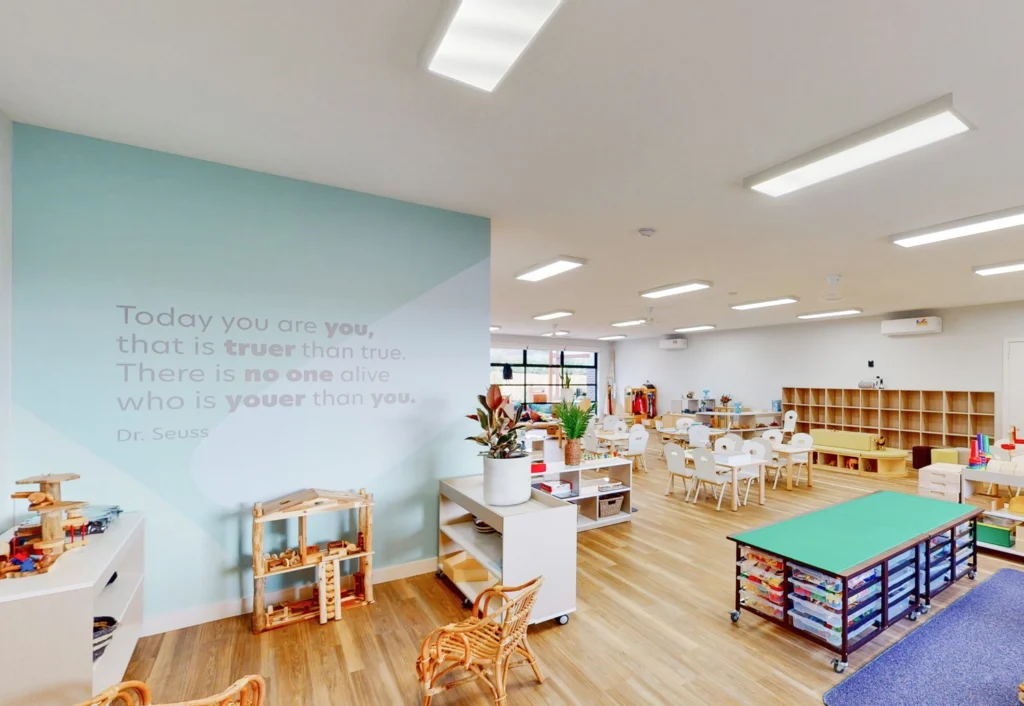 A brightly lit classroom with a Dr. Seuss quote on the wall, featuring tables, chairs, storage shelves, and various educational materials and toys.