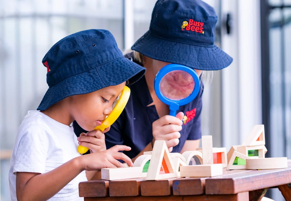 Two children in navy blue hats use magnifying glasses to examine wooden geometric shapes on a table.