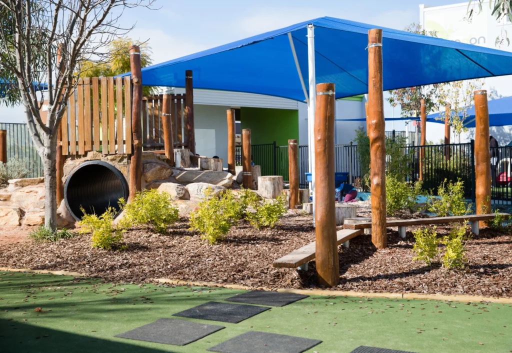 A children's playground with wooden structures, a tunnel, and a balance beam, shaded by a blue canopy. There are small bushes and a green rubber safety surface.