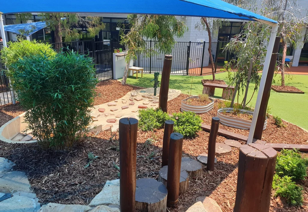A children's outdoor play area with stepping logs, a sandpit, shrubs, and a shaded area under a blue canopy, surrounded by a fence.