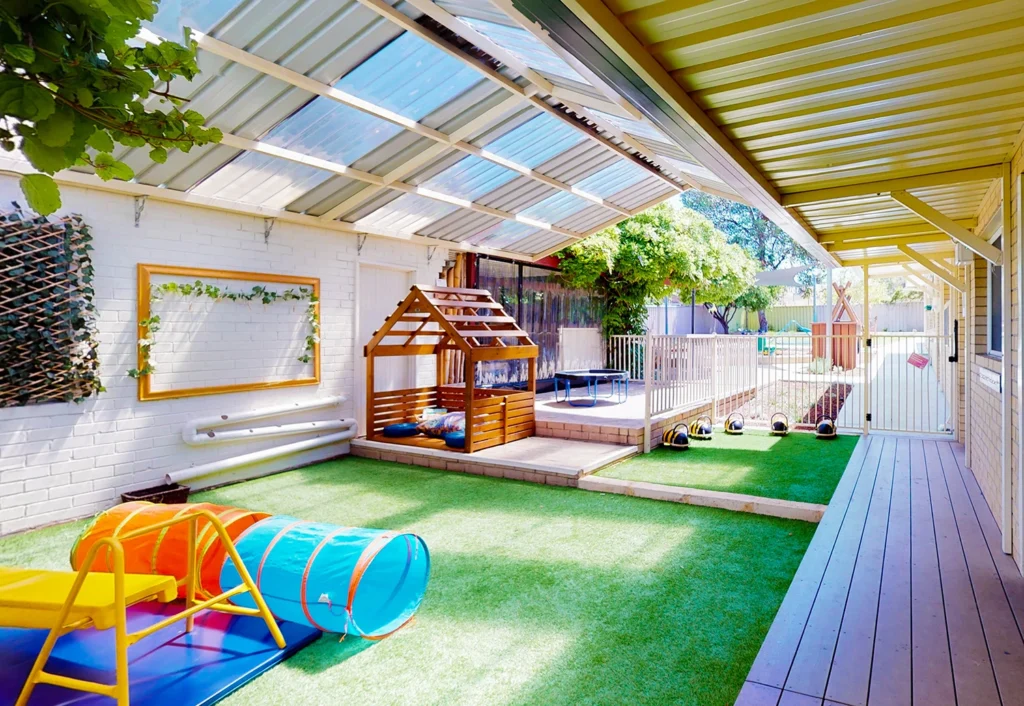 Outdoor children's play area with artificial grass, slide, tunnel, and small wooden playhouse. Overhead, a transparent roof provides shade. A fenced section in the background appears to contain a sandbox.