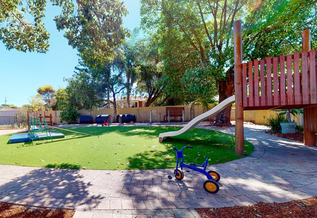 Outdoor playground with artificial grass, featuring a slide, a small climbing structure, tricycle, and large tires. The area is surrounded by trees and enclosed with a wooden fence.