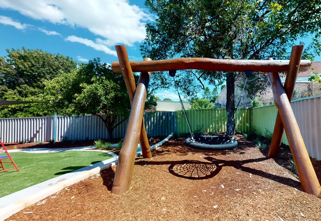 A backyard with a wooden swing set featuring a circular swing. The area has mulch ground cover, greenery, trees, a lawn, and a corrugated metal fence.