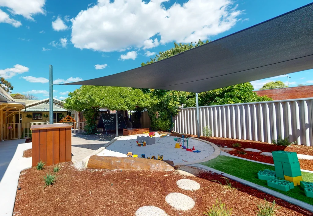A children's playground with a sandbox, play structures, and shaded areas. The ground is covered with mulch and stepping stones, while the sky is clear and blue with a few scattered clouds.