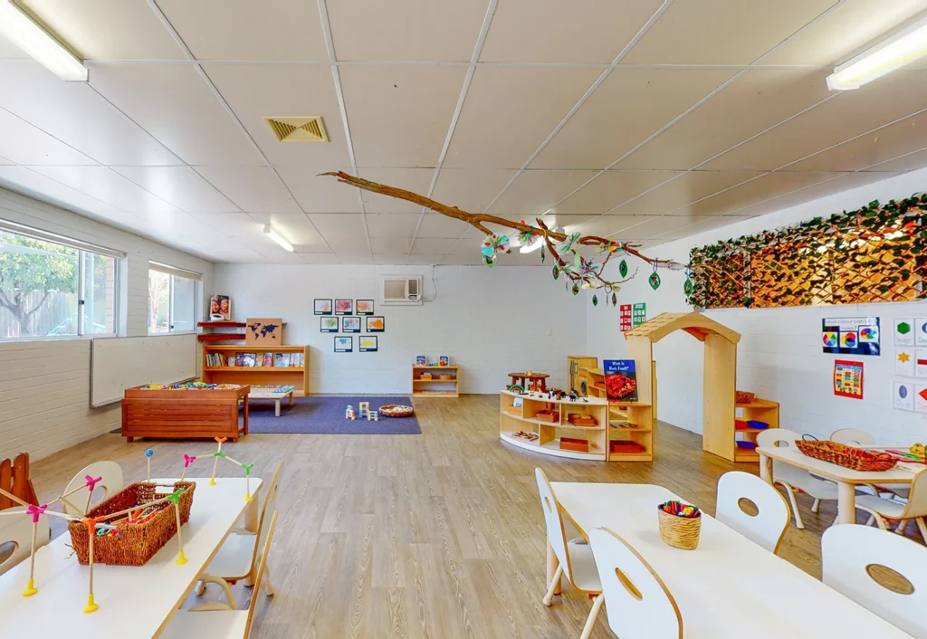 A brightly lit preschool classroom with tables, chairs, bookshelves, toys, and colorful decorations, including an overhead branch adorned with leaves and ornaments. Large windows allow natural light in.