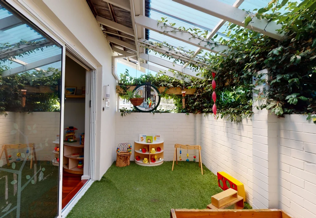 A covered outdoor play area with artificial grass, toys, and a hanging chair. The space includes a small shelf with books and toys. Sliding doors provide access to an adjoining room.
