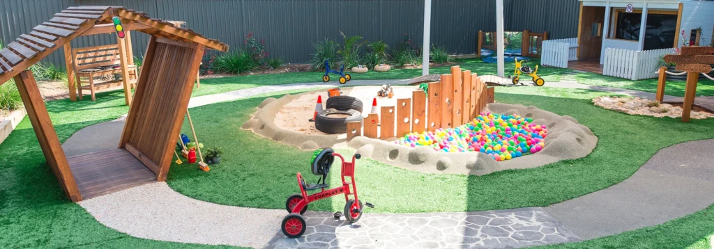 Outdoor playground featuring a colorful ball pit, tricycles, a wooden structure, and various play equipment on artificial grass and pebble pathways.