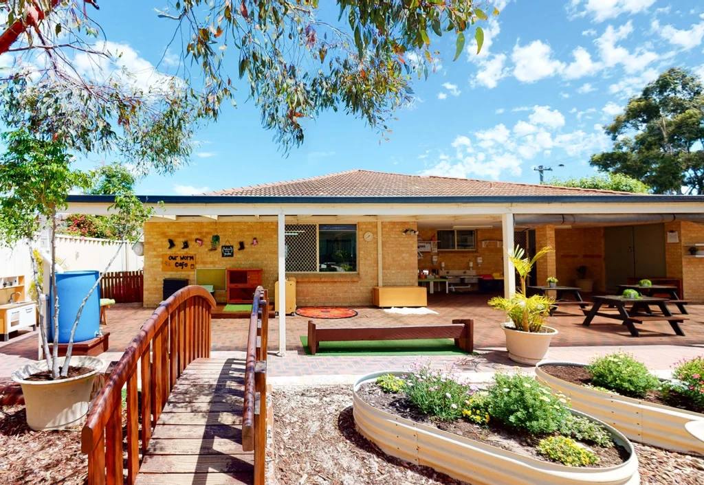 A view of a kindergarten's outdoor area features a wooden bridge, garden beds, picnic table, shade structure, and a building with a covered patio.