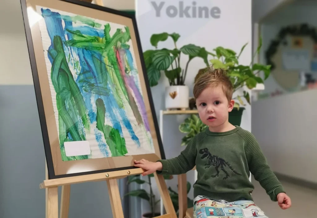 A young child in a green shirt stands beside an abstract painting on an easel, with various green plants in the background, capturing a creative moment at the day care.