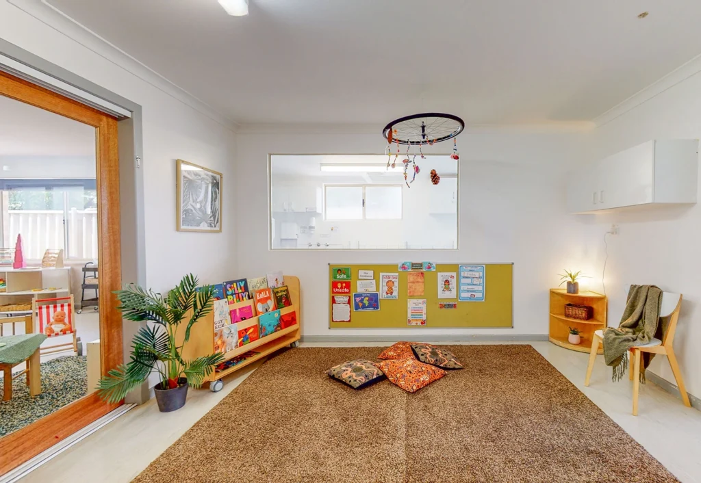 A children’s playroom with cushions on a brown rug, a dreamcatcher hanging from the ceiling, educational posters on the wall, and a bookshelf filled with books designed for early learning. This inviting space is perfect for kindergarten activities, fostering growth and creativity. A door leads to another room.