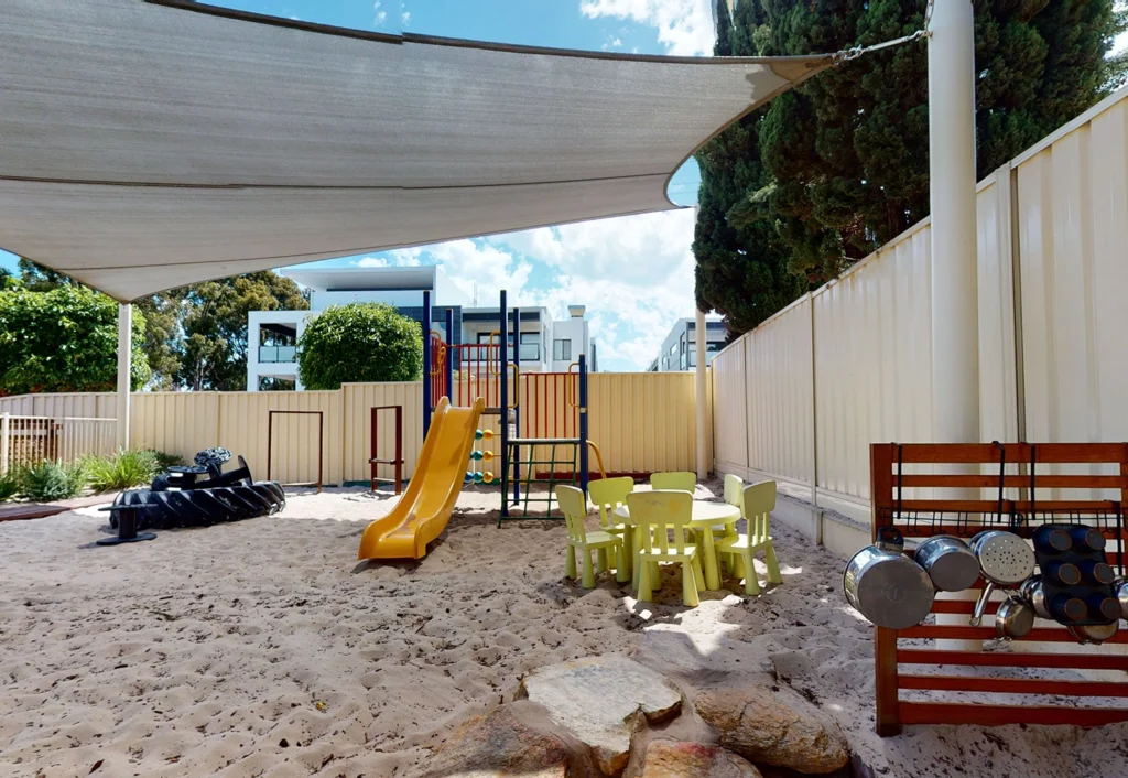 A preschool playground with a slide, climbing structures, small table and chairs set, and a play kitchen area with pots and pans. The ground is covered with sand, and a shade cloth is overhead—perfect for childcare activities.