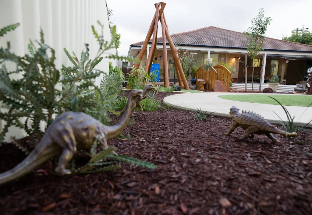 Outdoor scene featuring a playground with wooden structures, greenery, and dinosaur figurines placed among mulch. A house with a tiled roof is in the background, ideal for an early learning daycare setting.
