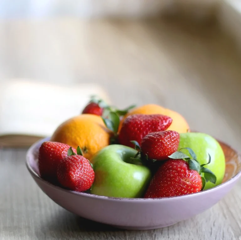A bowl of fresh fruits including green apples, strawberries, and oranges on a wooden table, reminiscent of a vibrant kindergarten snack time. In the background, a blurred open book adds to the charm of this early learning setting.