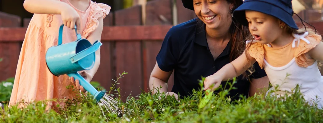 Two young girls, wearing hats and aprons, water plants in a garden bed while a woman kneels beside them, smiling and assisting. It's a delightful moment of early learning and childcare in nature's classroom.