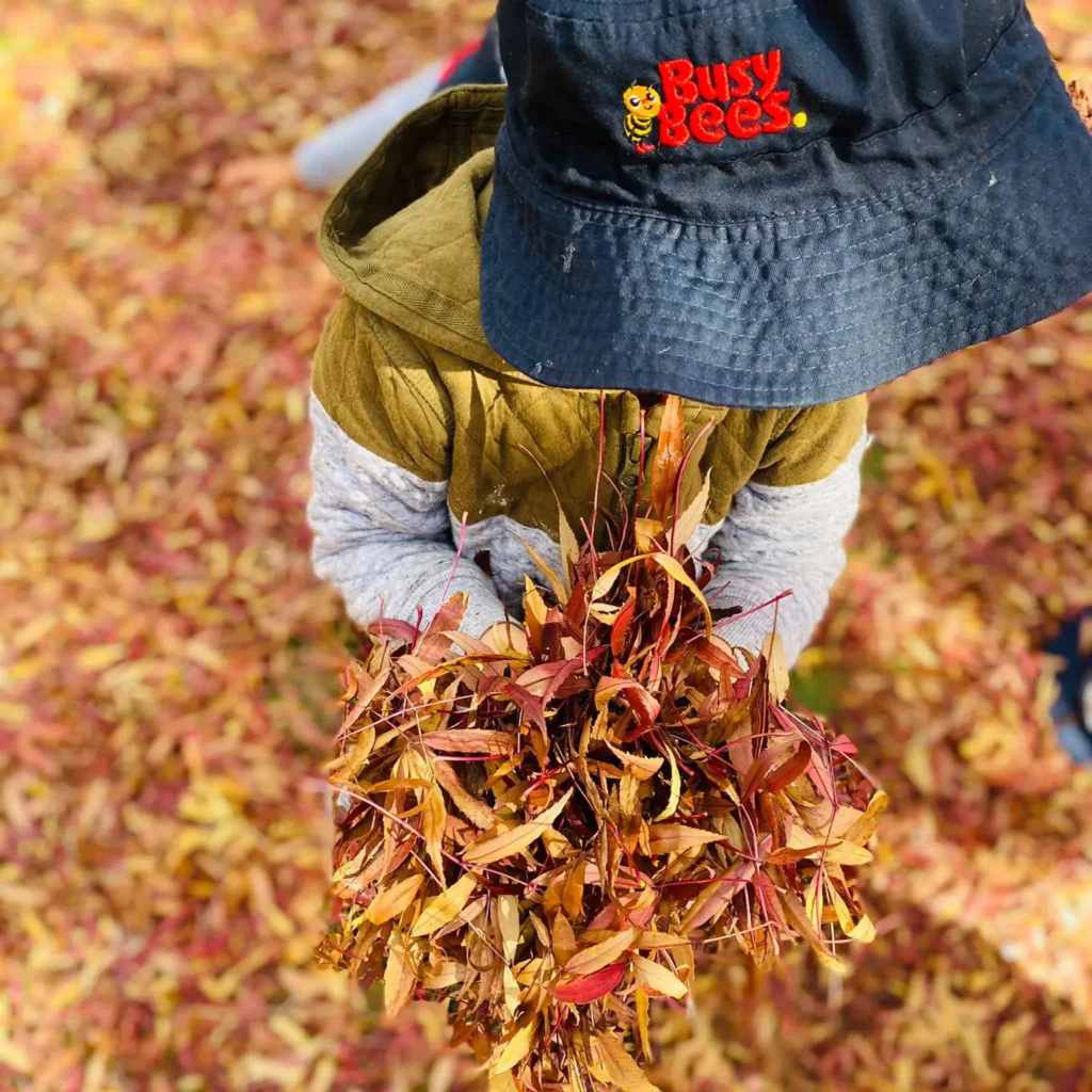 A child from the Busy Bees childcare center holds a bundle of autumn leaves while wearing a black hat with the "Busy Bees" logo and a green jacket.